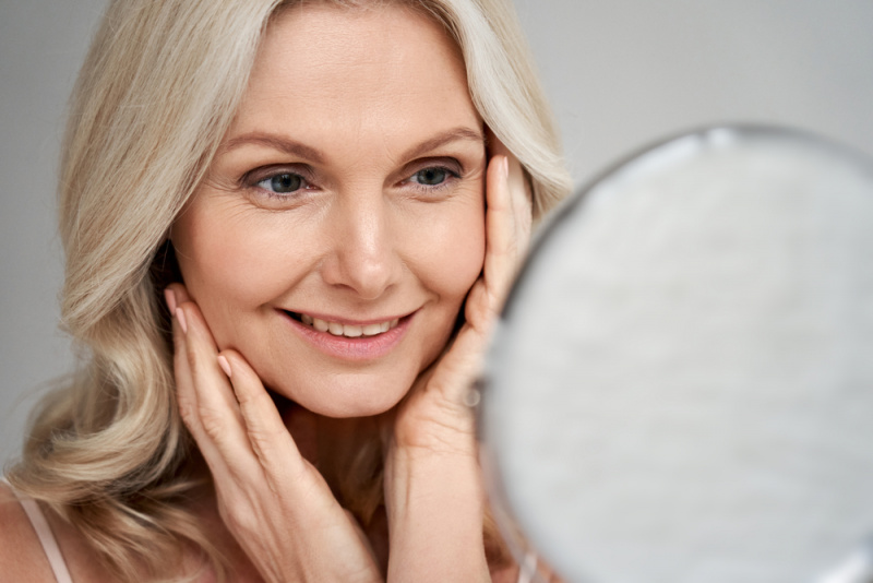 bella donna anziana capelli biondi si specchia allo specchio con mani sul viso pelle