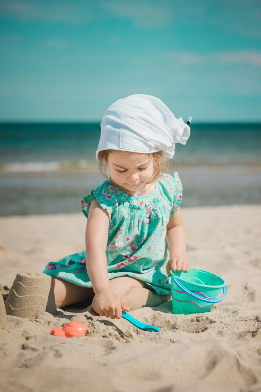 bella bambina abito leggero turchese con fantasia a fiori cappello bandana sui capelli biondi gioca con palette e secchiello sulla spiaggia al mare