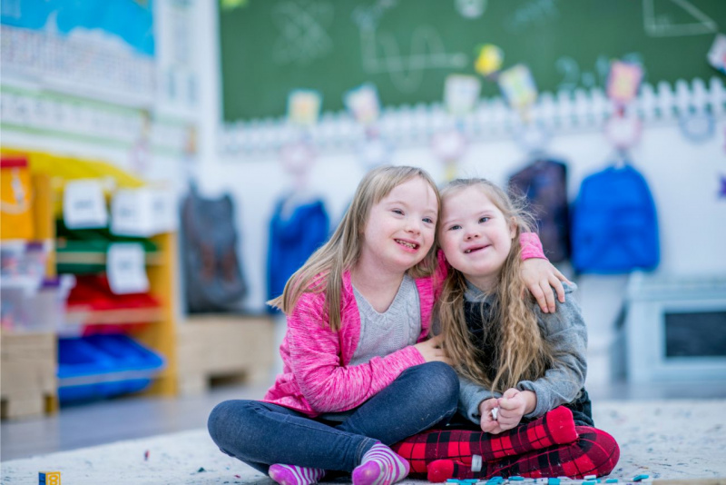 due belle bambine capelli lunghi biondi sorridono e si abbracciano sedute all'asilo scuola materna inclusione sindrome di Down