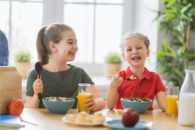 colazione al mattino tazze cereali bambine cucchiaio in mano mela bottiglia latte bicchiere succo arancia