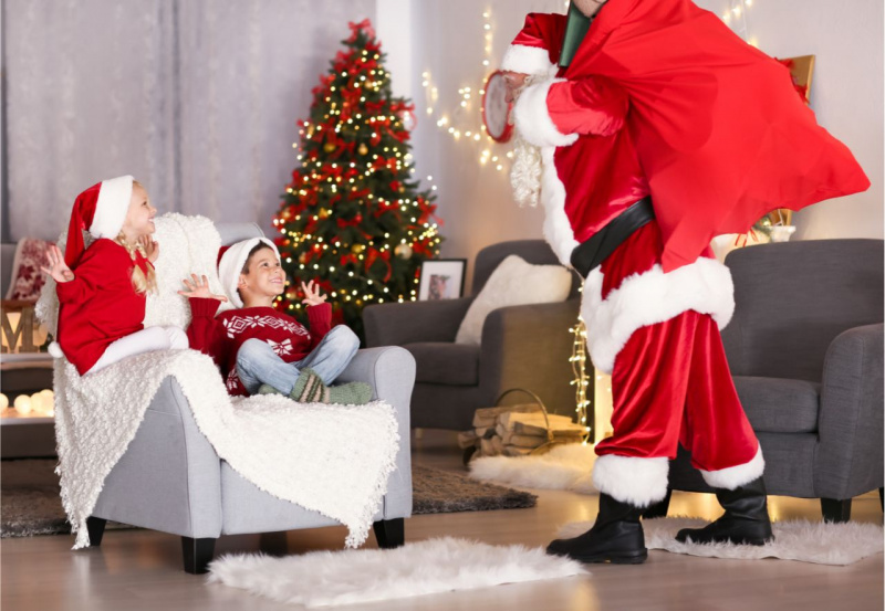 bambina trecce bionde bambino sorriso cappello rosso bianco seduti su poltrona guardano felici Babbo Natale sacca rossa davanti Albero luci decorazioni natalizie regali