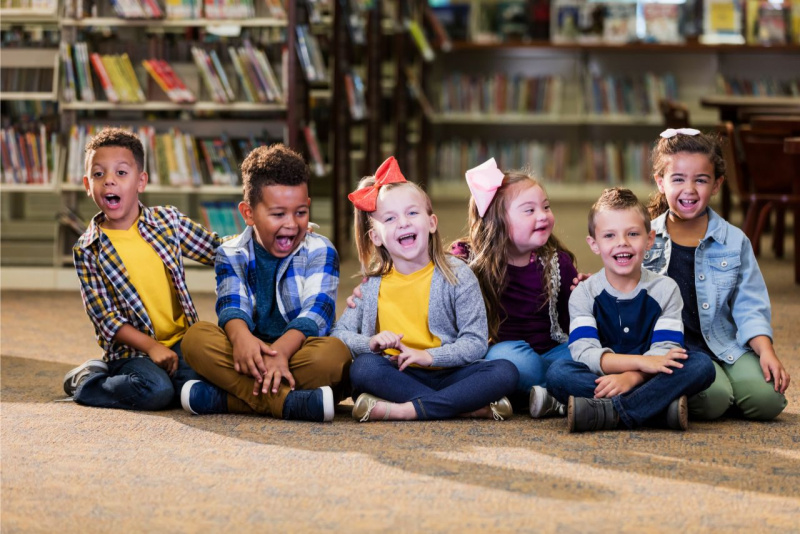 bellissimi bambini seduti per terra in blblioteca ridono e si divertono inclusione sindrome di down