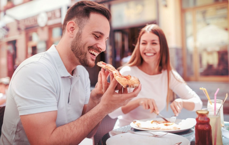 uomo bello mangia fetta di pizza con sorriso mentre la donna lo guarda pizzeria all'aperto sorrisi risate