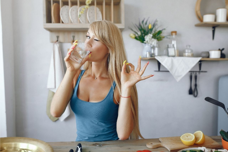 giovane bella donna capelli biondi beve un bicchiere d'acqua in cucina occhiolino ok