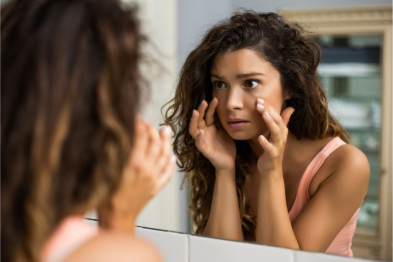 occhiaie segni di stanchezza bella donna capelli lunghi ricci castani si tocca viso guardandosi allo specchio canotta rosa