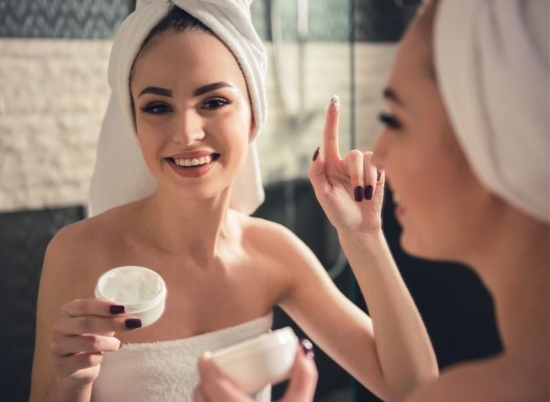 giovane bella donna applica crema viso davanti lo specchio bagno