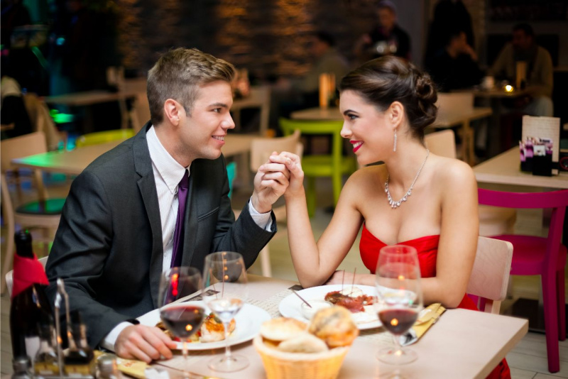 uomo affascinante tiene mano bella donna abito rosso seduti a tavola ristorante bottiglia vino rosso calici cesto pane piatti