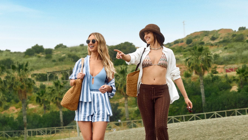 Due giovani donne sorridenti indossano una il bikini con sopra camicia bianca, pantaloni marrone pizzo, l'altra costume intero celeste con sopra completo camicia shorts righe bianco azzurro,borsa paglia, cappello occhiali da sole, rappresentando la moda mare e i costumi da bagno da donna dell'estate.