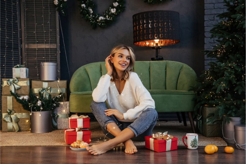 Giovane donna in jeans e maglione bianco seduta sul pavimento di casa circondata da pacchi regalo, lucine led e un albero di Natale illuminato.