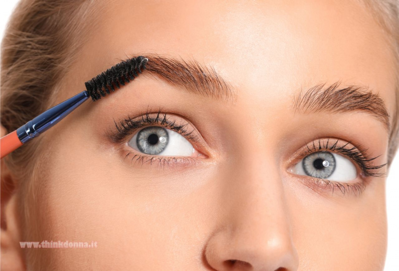 Primo piano di una giovane donna bionda con occhi azzurri e un trucco naturale, che si pettina le sopracciglia verso l'alto con un pettinino scovolino per dar loro ordine e forma.