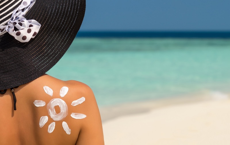 protezione solare donna di spalle con crema solare a forma di sole sulla spalla cappello nero fiocco pois bianco nero mare estate 