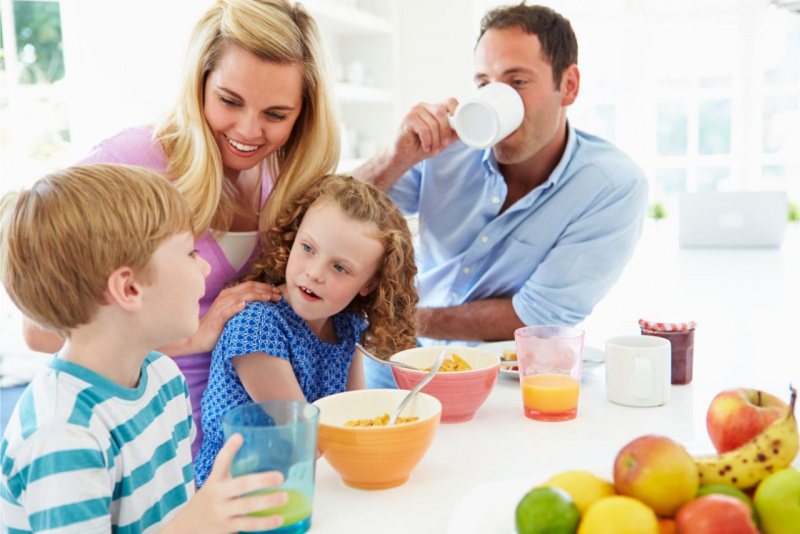 fare colazione insieme famiglia allegra mamma donna capelli biondi sorriso figlio bambino con bicchiere in mano succo arancia bambina figlia che lo guarda ciotole cereali uomo pap beve latte confettura frutta barattolo