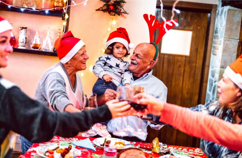 Famiglia multigenerazionale festeggia il Natale a tavola con il nonno che tiene in braccio la nipotina e la nonna in grembiule che sorride.