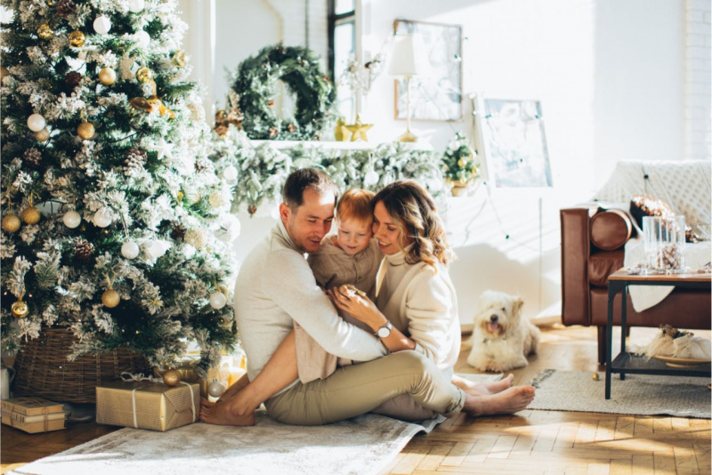 albero di Natale decorato oro bianco cesta vimini pacchi regalo libri uomo papà abbraccia donna mamma e figlio piccolo soggiorno divano pelle