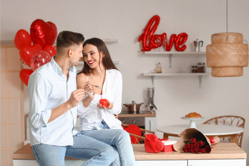 bella donna sorridente guarda uomo mentre dividono dolce da mangiare insieme scritta love palloncini rosso rose regalo