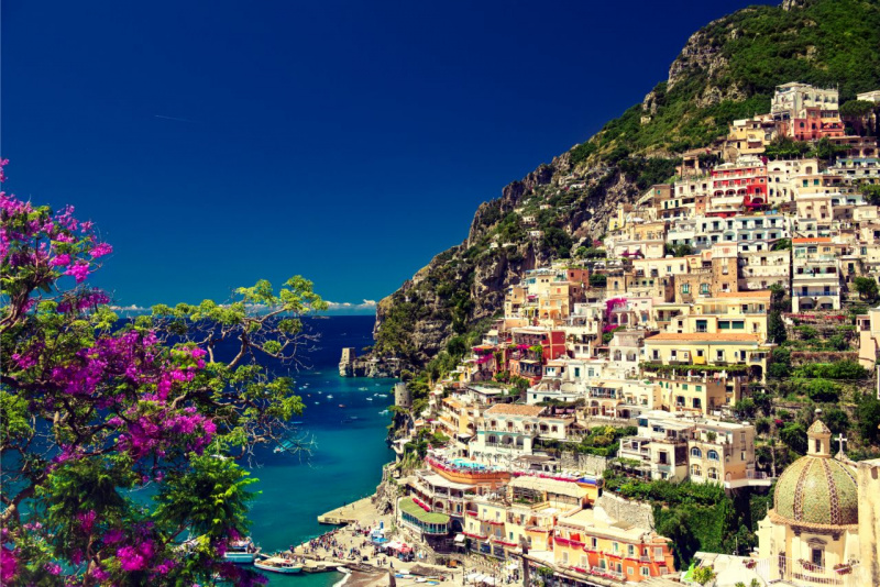 Scorcio panoramico di Positano con fiori fucsia in primo piano, le case colorate sulla scogliera e il mare blu turchese