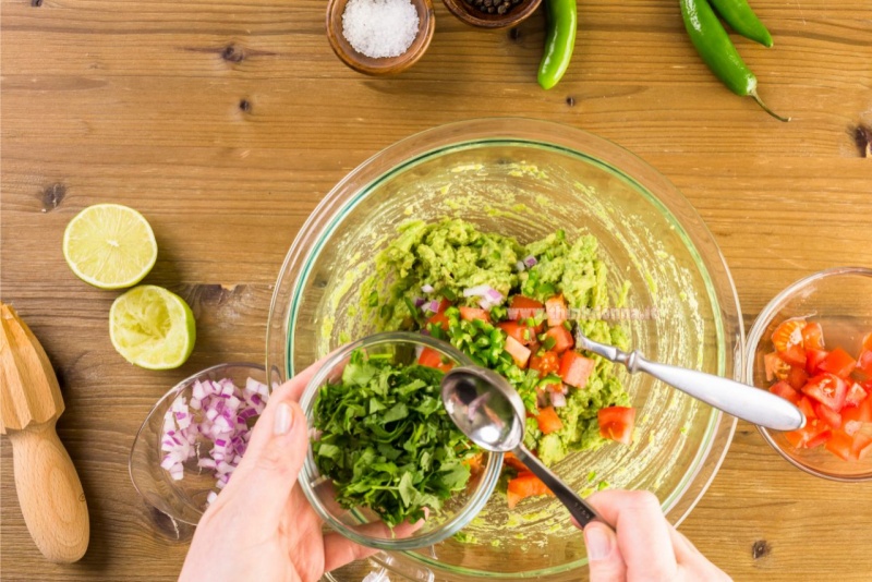 ingredienti preparazione guacamole polpa di avocado foglie coriandolo pezzi pomodoro rosso cipolla tritata peperoncino verde piccante cucchiaio ciotola vetro su tavolo legno