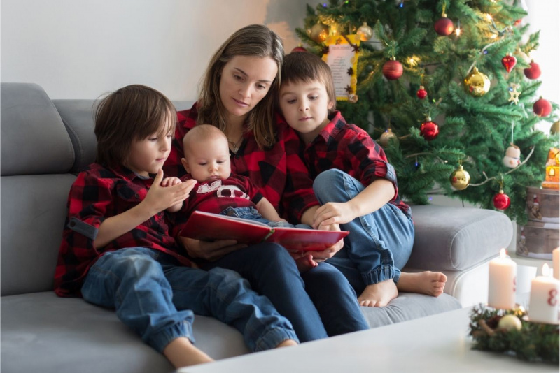 madre con figli piccoli seduti sul divano leggono libro con albero di natale 