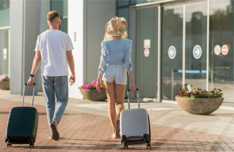uomo jeans t-shiert bianca di spalle cammina con donna shorts cappello camicia verso entrata aeroporto con trolley viaggi